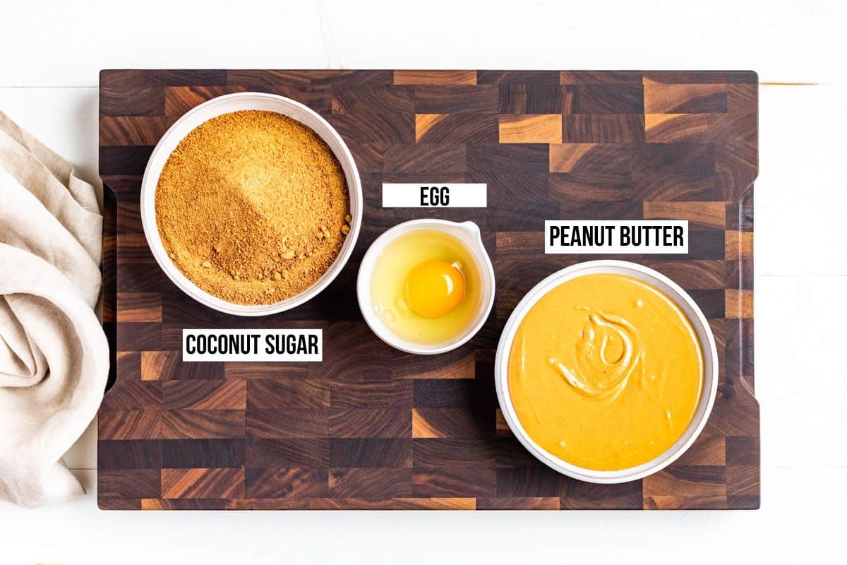 Peanut butter, coconut sugar, and an egg in bowls on a wood cutting board.