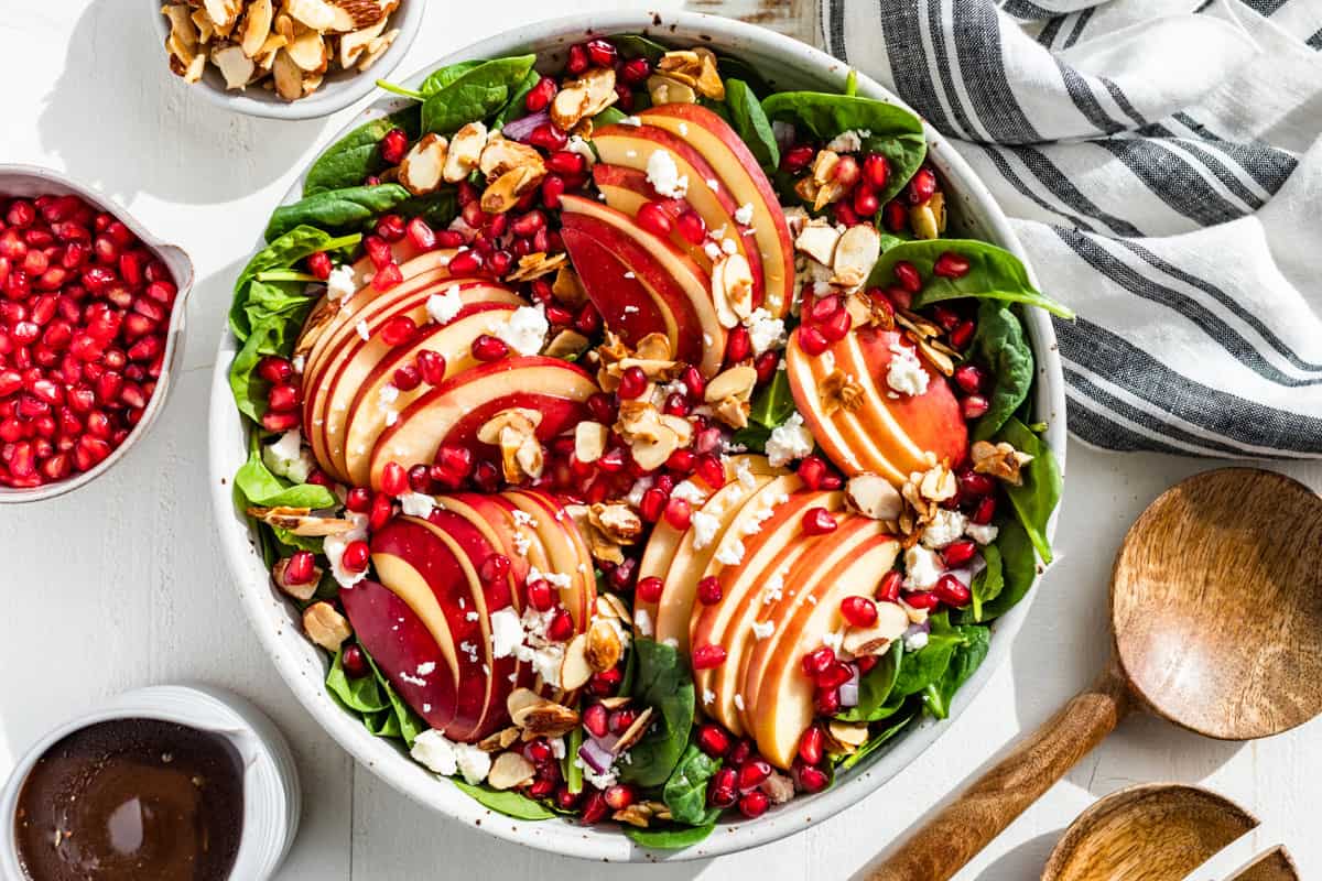 Apple Spinach Salad being layered into the serving bowl with pomegranate arils, balsamic vinaigrette, and almonds on the side.