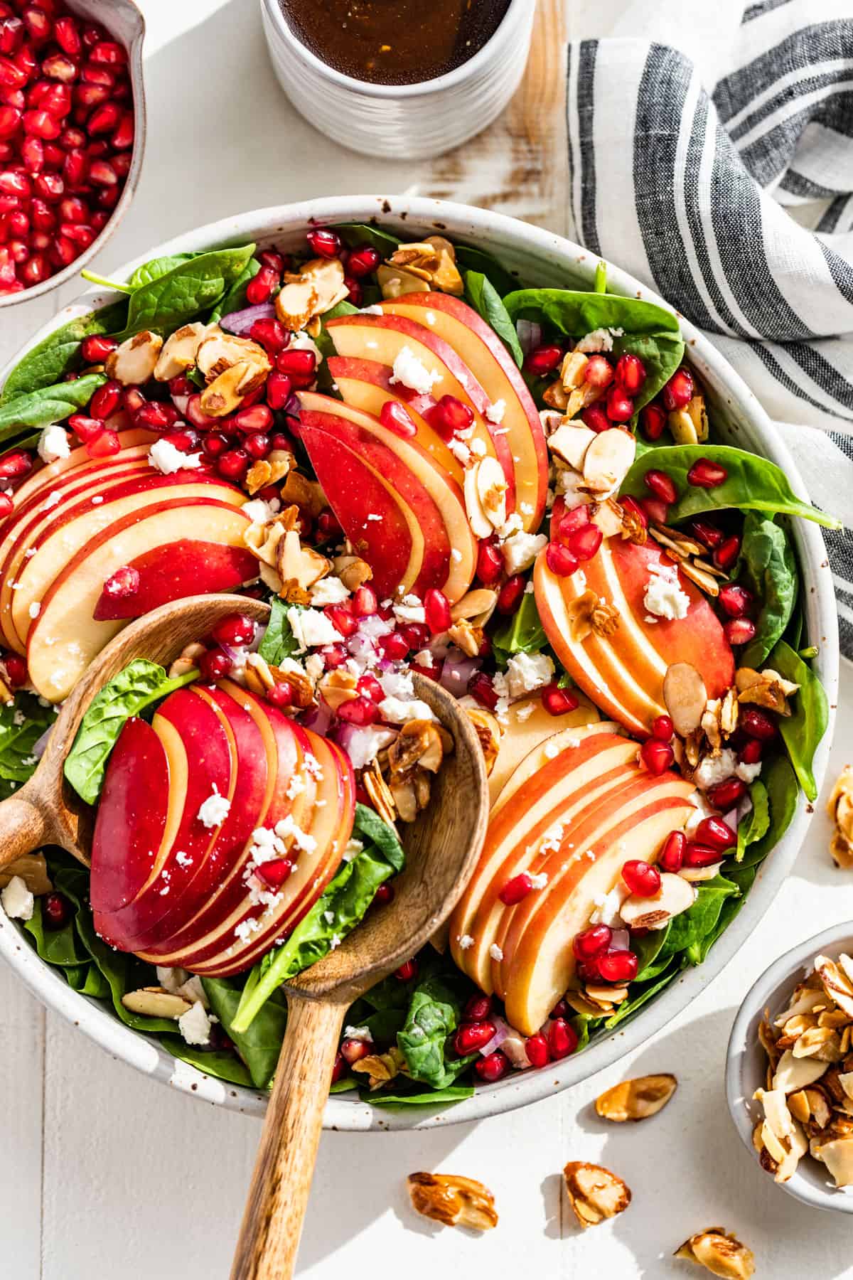 Straight down view of Apple Spinach Salad in a pottery bowl with wooden serving spoons and balsamic vinaigrette & pomegranate arils on the side.