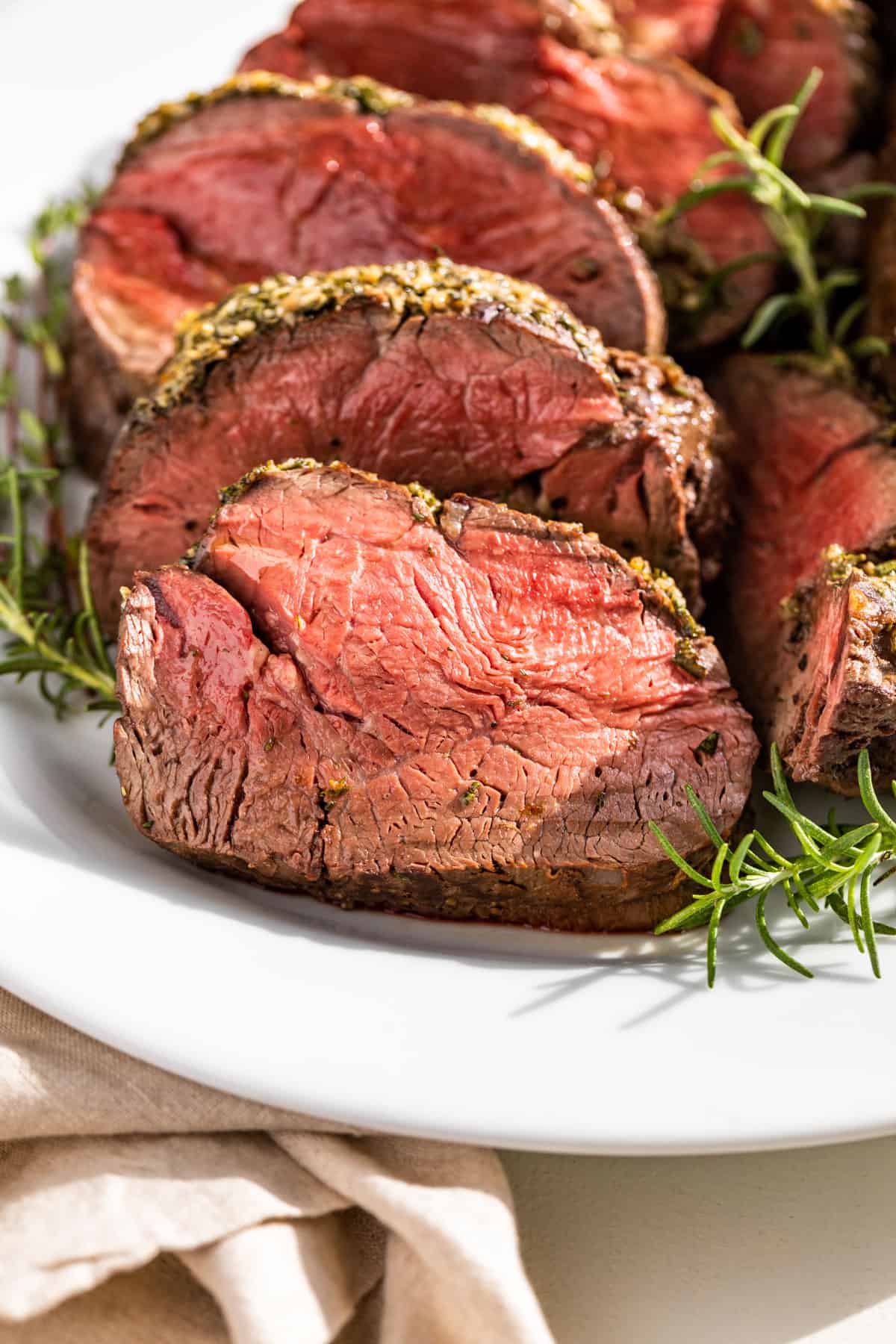 Side view of sliced Beef Tenderloin on a white serving platter with rosemary sprigs around it.