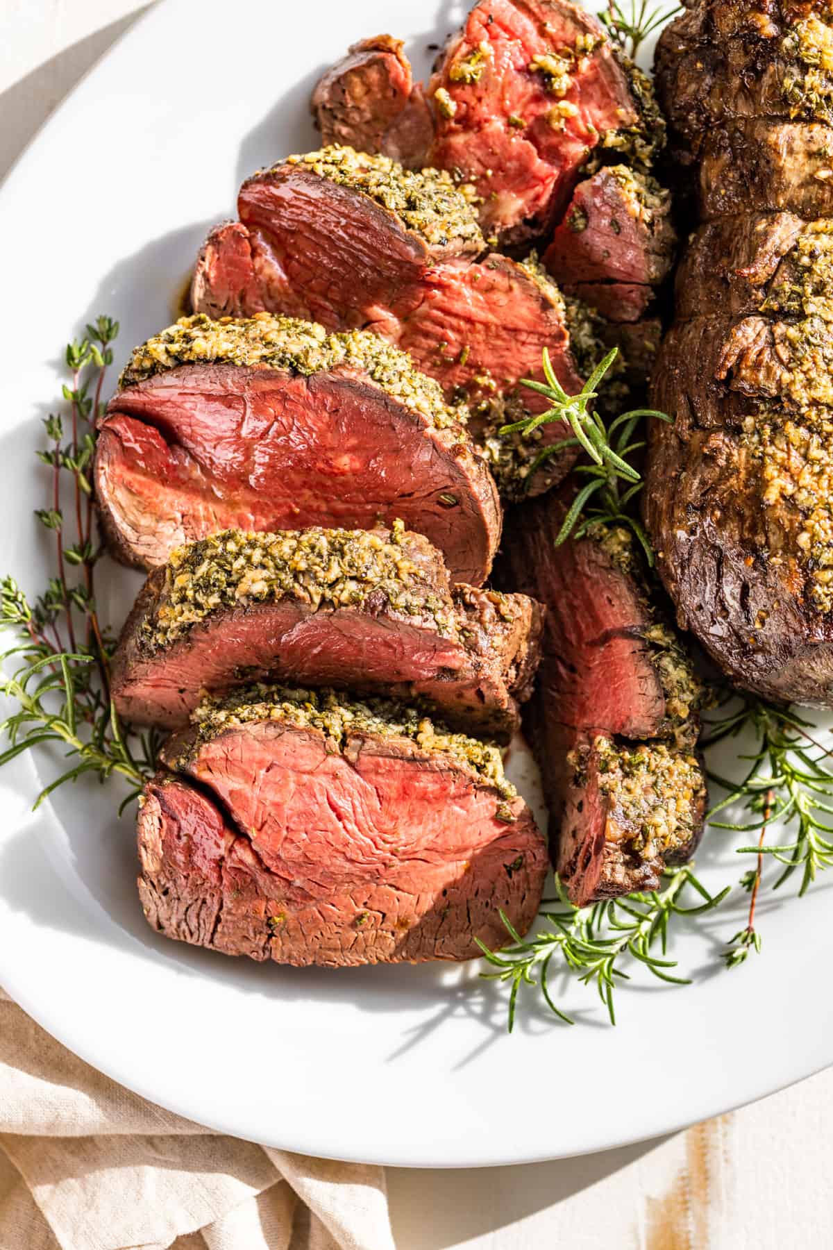 Downwards view of sliced roast Beef Tenderloin with fresh rosemary and thyme sprigs around it.