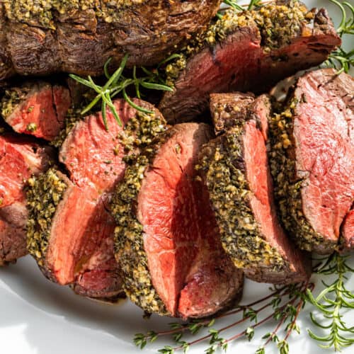 Sliced Beef Tenderloin on a white serving platter surrounded by sprigs of thyme and rosemary.