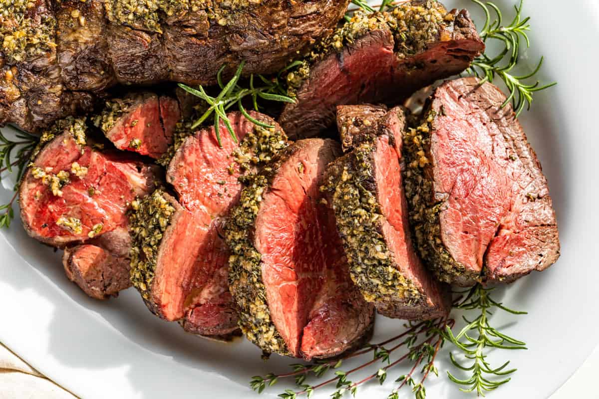 Sliced Beef Tenderloin on a white serving platter surrounded by sprigs of thyme and rosemary.