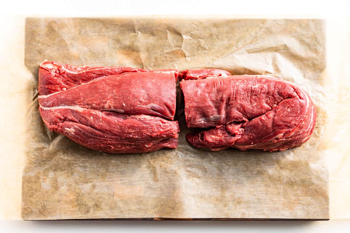 The first step of cutting the whole beef tenderloin in half on brown paper on a wood cutting board.
