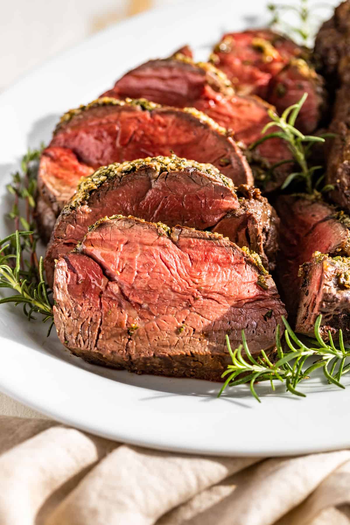 Sliced Roast Beef Tenderloin on a white plate surrounded by rosemary and thyme sprigs.