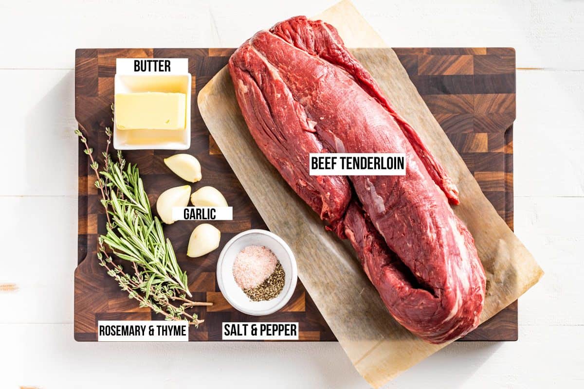 Beef Tenderloin ingredients on a wood cutting board, a whole beef tenderloin, butter, thyme, rosemary, garlic, sea salt and pepper.