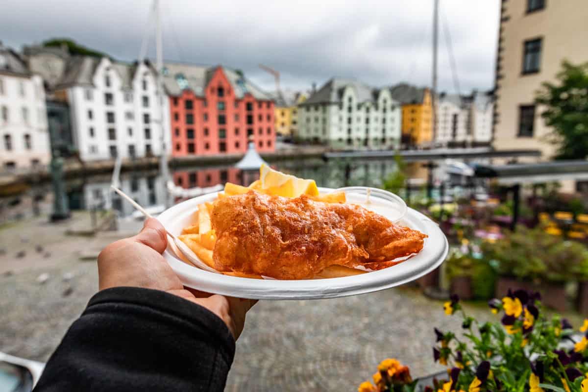 A hand holding a plate of fish and chips with waterfront housing in the background.