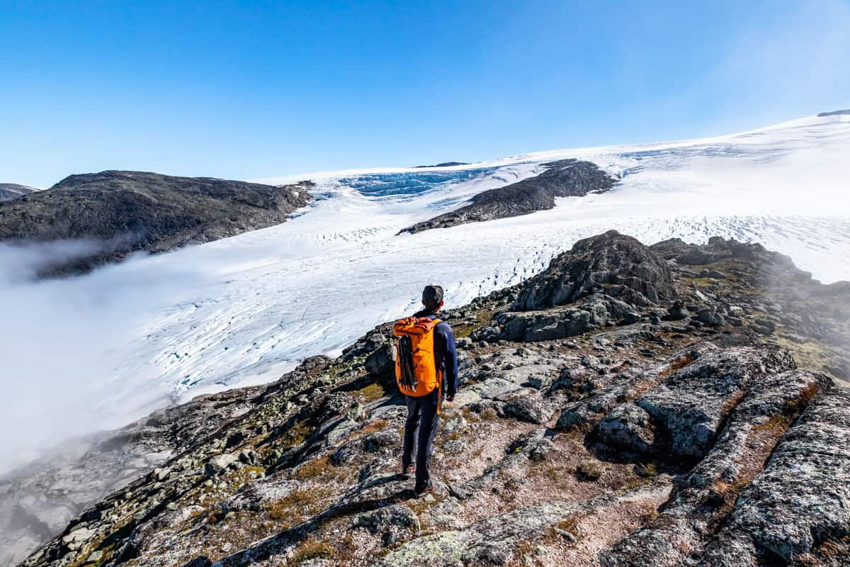 Man with an orange backpack standing on a rocky mountain top overlooking a vast glacier with some fog around the edges.
