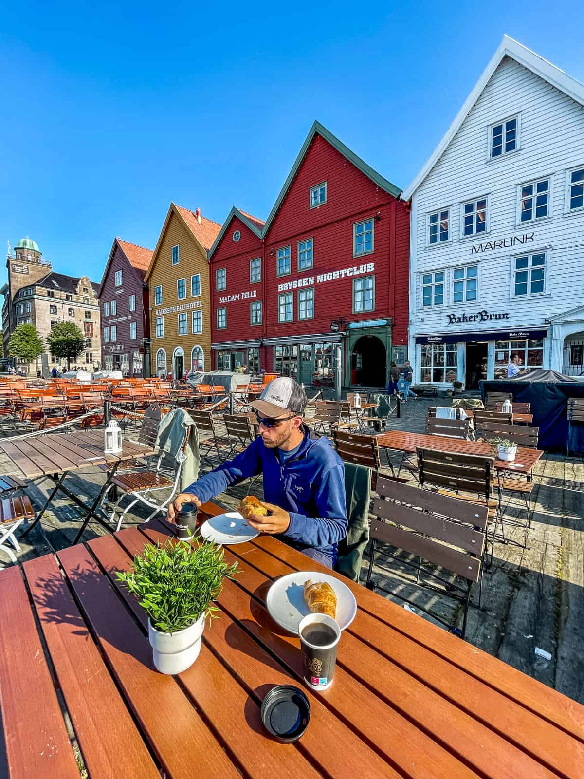 Man at an outdoor table eating a croissant in front of the old fishing houses called Bryggen a world Unesco heritage sight.
