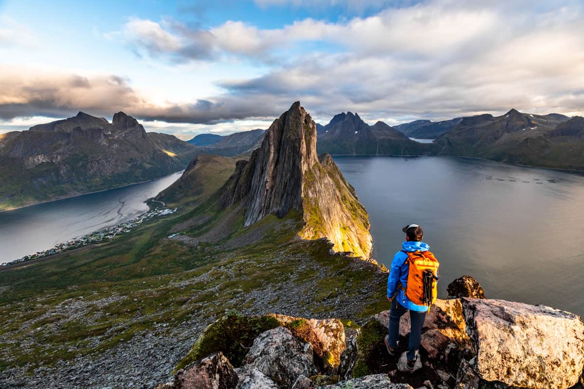 Man with an orange backpack overlooking Segla mountain with the ocean surrounding it at sunset.