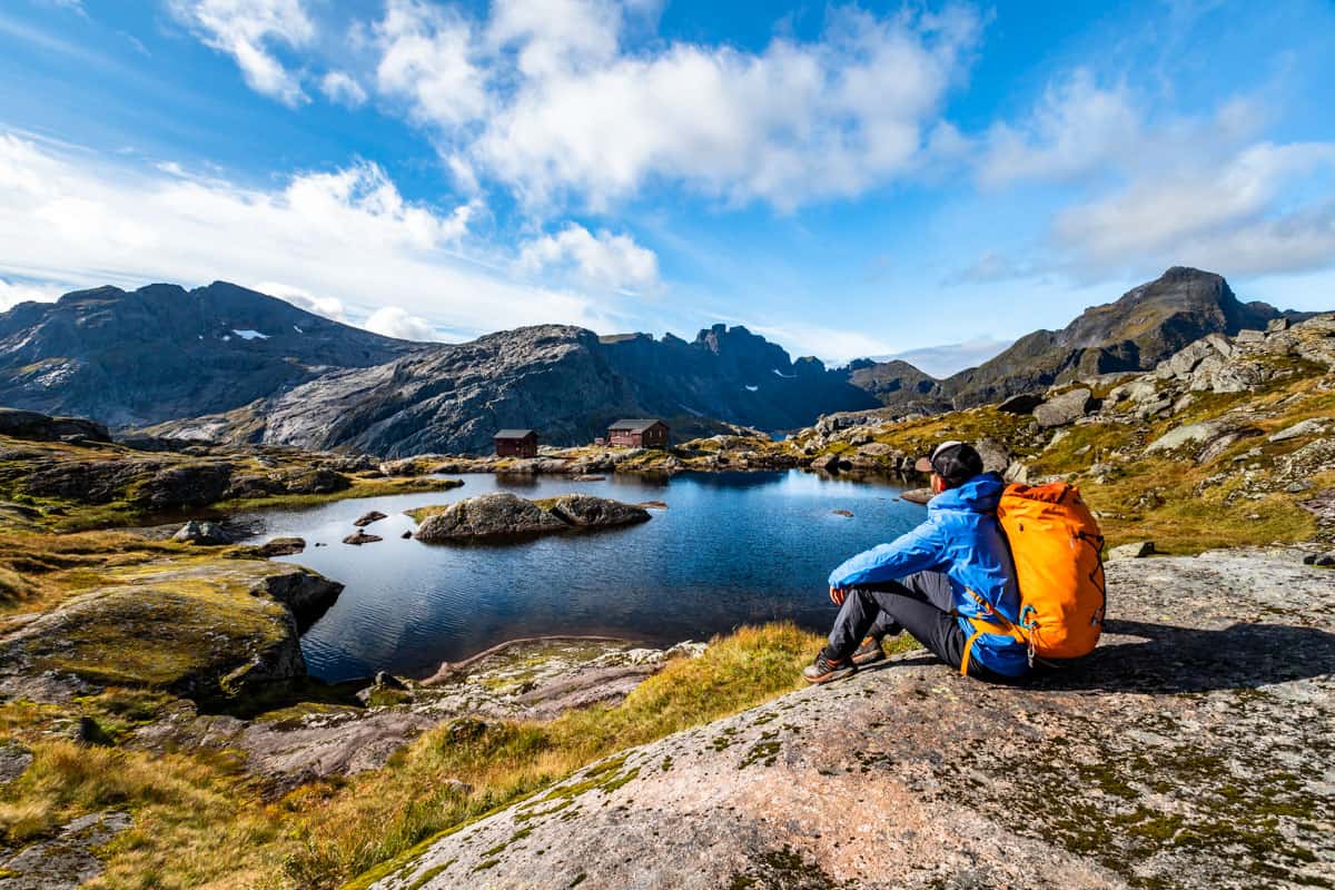 Man with an orange backpack sitting on a rock overlooking a pond in front of the Munkebu Hut, Norway.