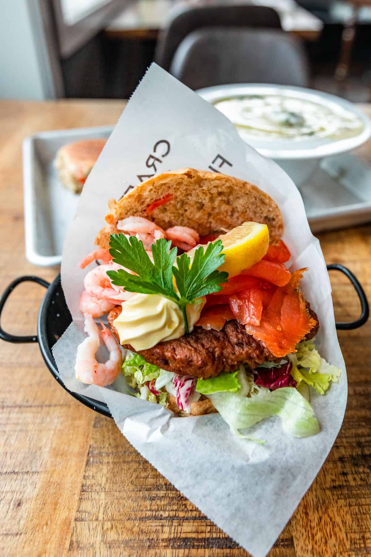 A fish burger topped with smoked salmon and shrimp in a black serving dish on a wood table.