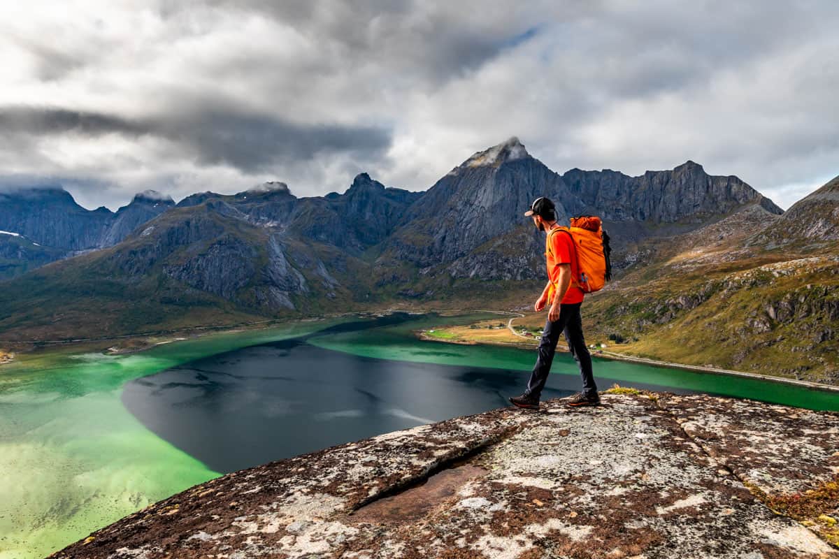 Man with an orange backpack walking on a rock slab on a mountain top overlooking the ocean with mountains in the background.