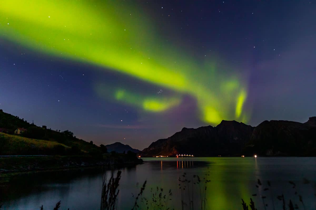 Green northern lights arcing across a dark sky with water in the foreground and mountains in the background.