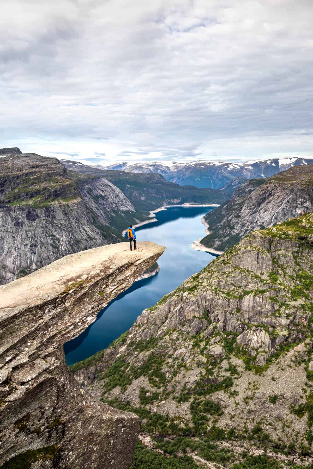 Man with an orange backpack standing on the 'trolls tongue' rock overhang on the Trolltunga hike in Norway looking over a fjord.