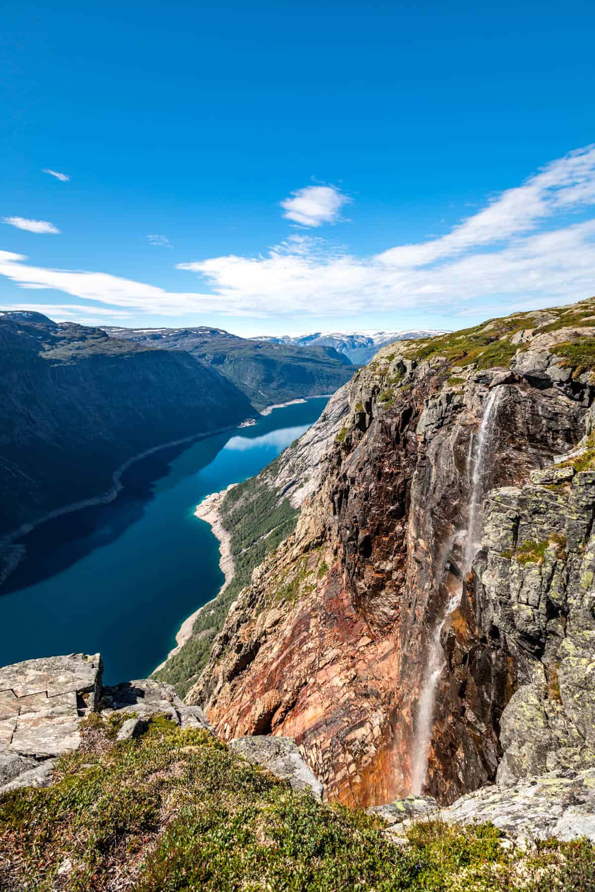 A waterfall pouring out of a steep hillside into the blue fjord below on a sunny day.