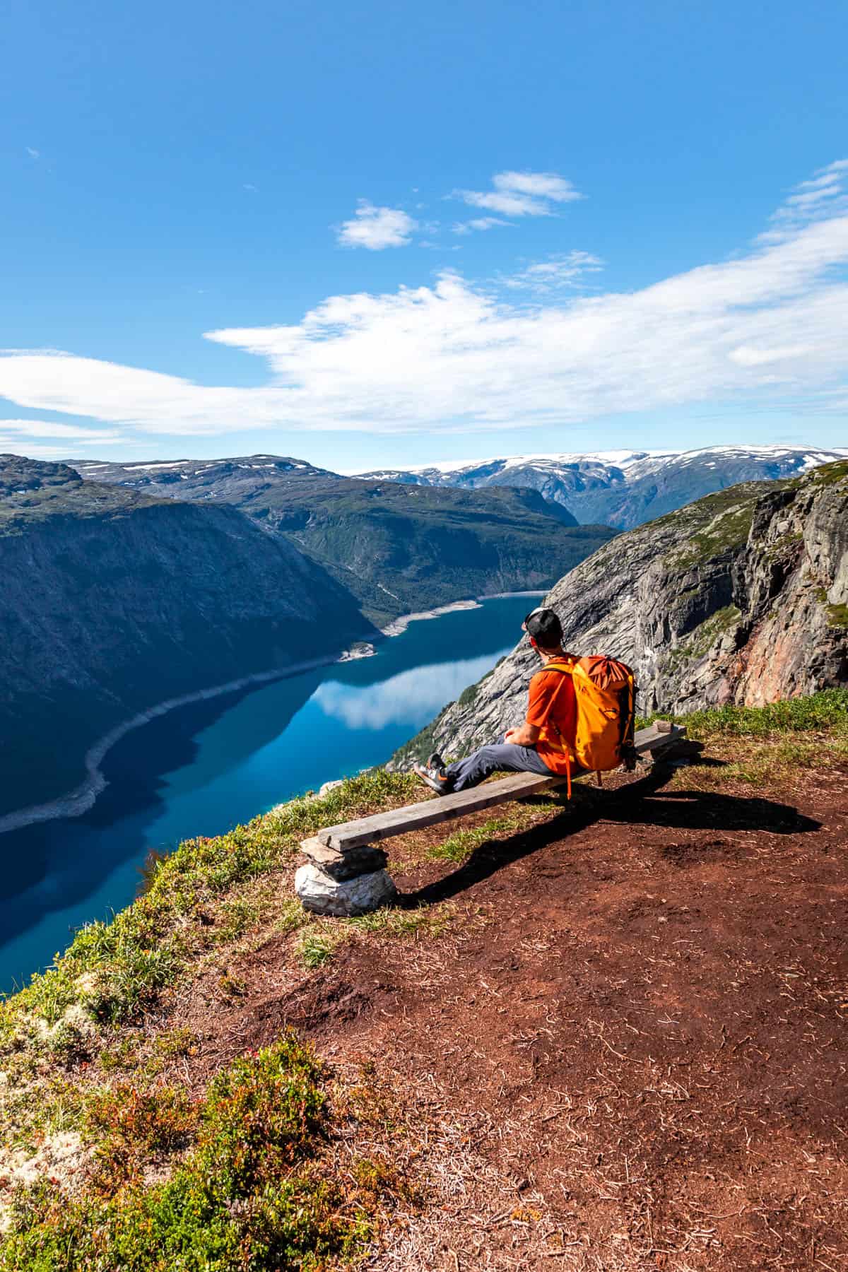Man with an orange backpack sitting on a low wooden bench overlooking a fjord with sunny blue skies.