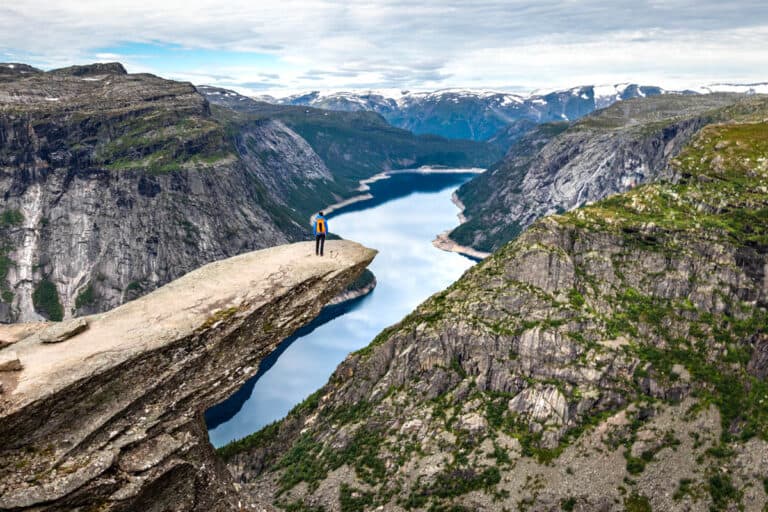 Man with an orange backpack standing on the rocky outcropping called the troll's tongue overlooking a fjord in Norway.