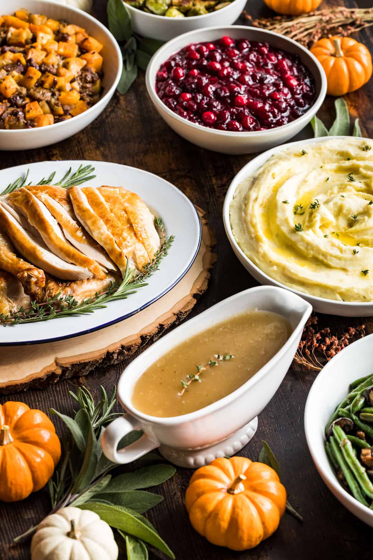 A wood table filled with a gravy boat, a platter of sliced turkey, mashed potatoes, cranberry sauce, butternut stuffing, and green beans surrounded by decorative pumpkins.