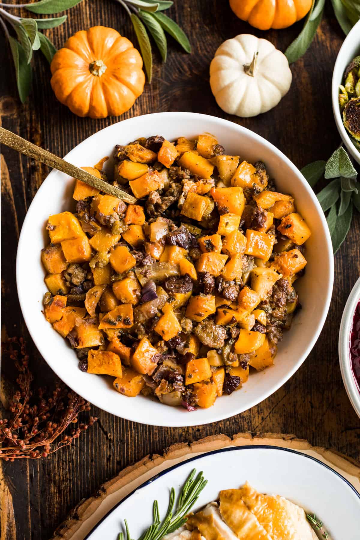 A bowl of butternut stuffing in the center surrounded by decorative pumpkins and a platter of sliced turkey.