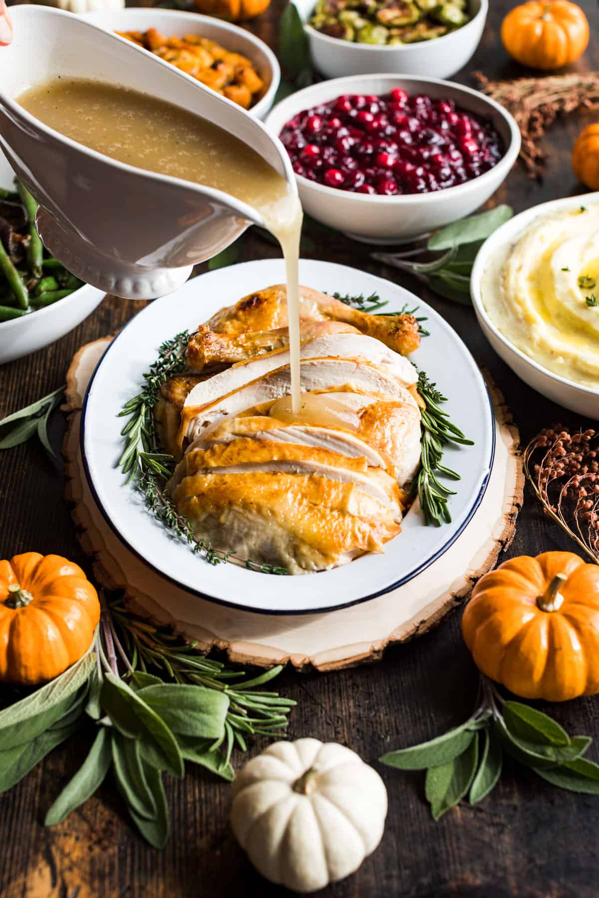 A gravy boat pouring gravy over a platter of sliced turkey with decorative pumpkins and Thanksgiving side dishes around it.