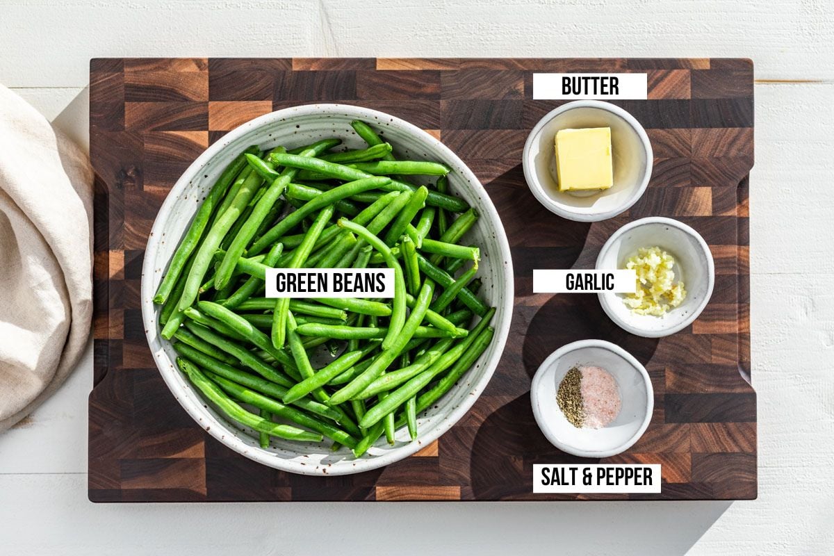 Raw green beans in a bowl with small bowls of butter, garlic, sea salt and pepper on a wood cutting board.