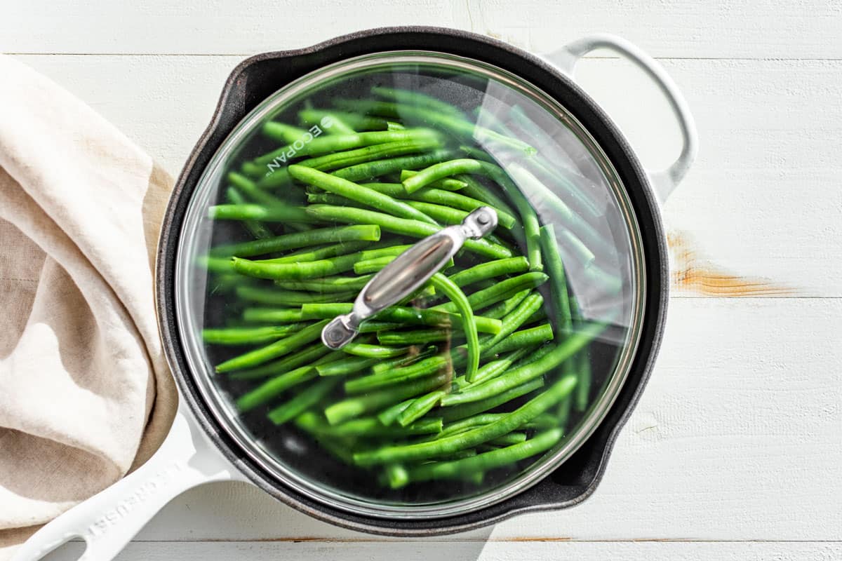 Green beans in a white cast iron skillet with a lid on top being steamed.