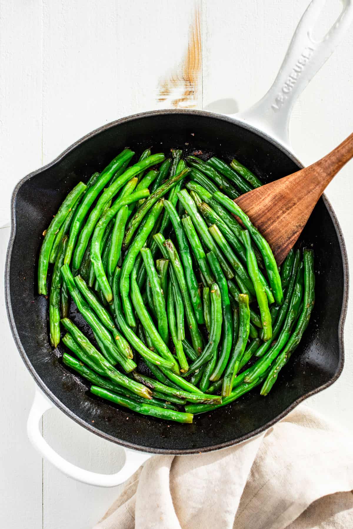 Downwards view of Sautéed Green Beans in a white skillet with a linen on the side and a wood spatula on the side of the skillet.