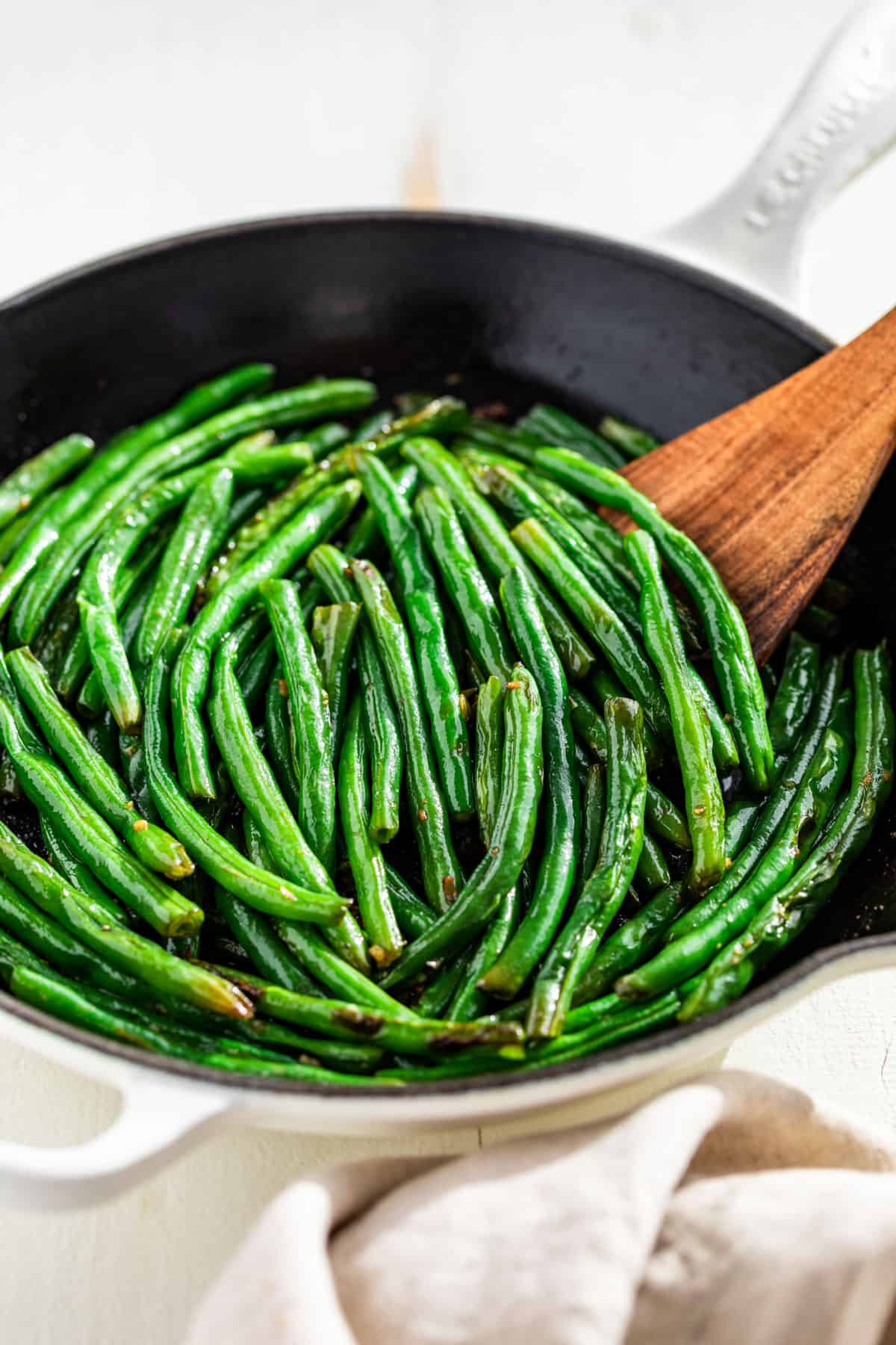 Side view of Sautéed Green Beans in a white skillet with a wood spatula on the side.