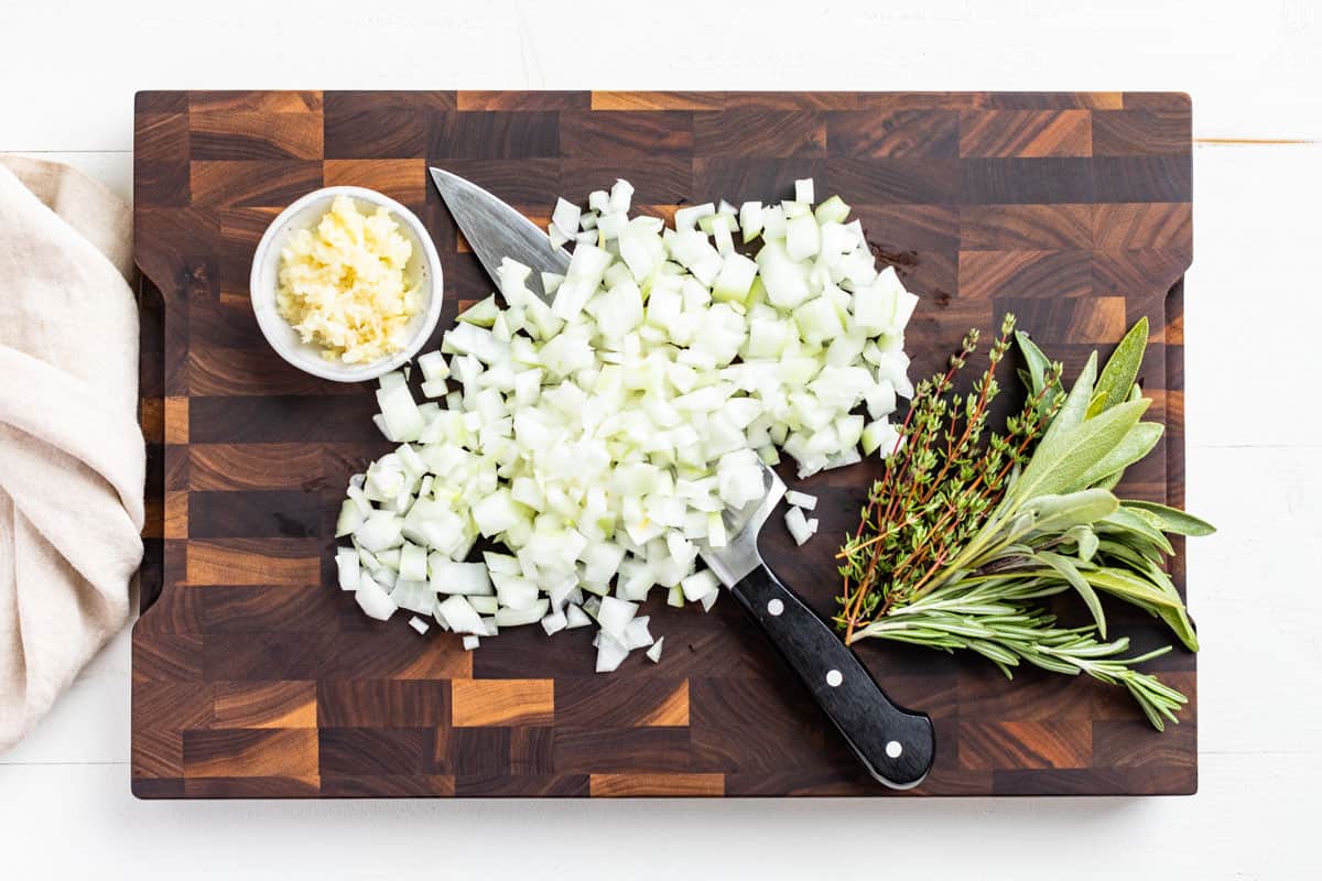 Diced onion, minced garlic, and fresh herbs on a wood cutting board.