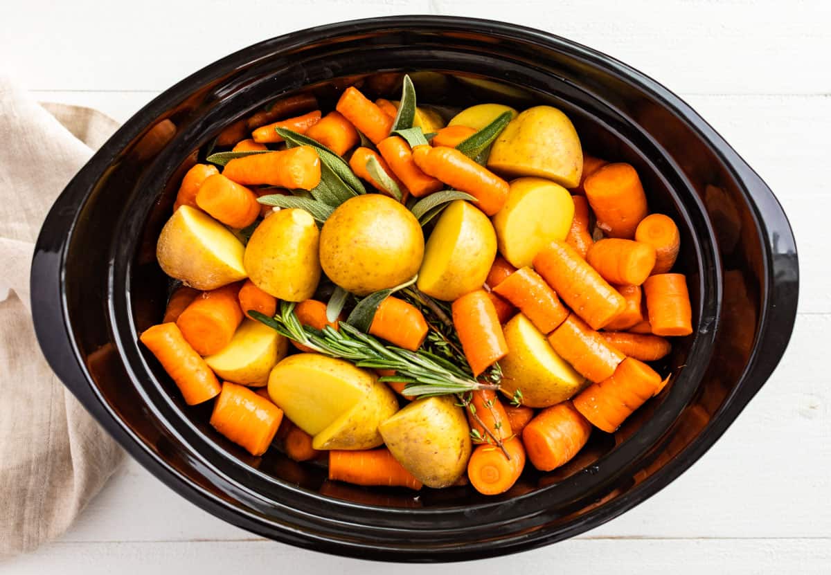 The potatoes, carrots, and fresh herbs being adding to the slow cooker bowl on top of the browned pork roast.