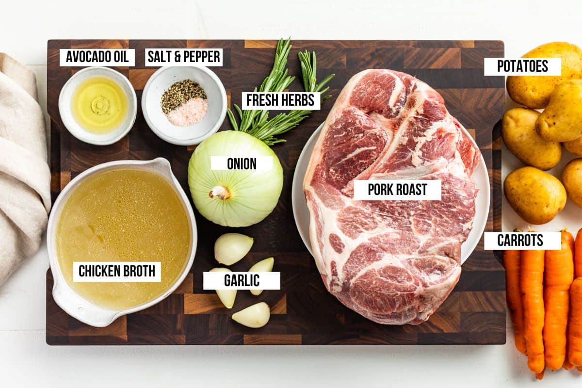 All the ingredients for Slow Cooker Pork Roast in containers on a wood cutting board, pork shoulder roast, carrots, potatoes, onion, fresh herbs, garlic, and chicken broth.