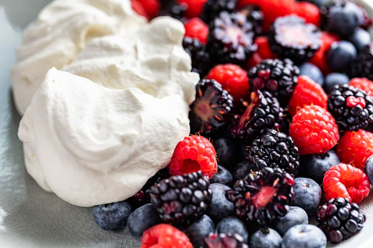 A dollop of whipped cream on a grey plate next to raspberries, blueberries, and blackberries.