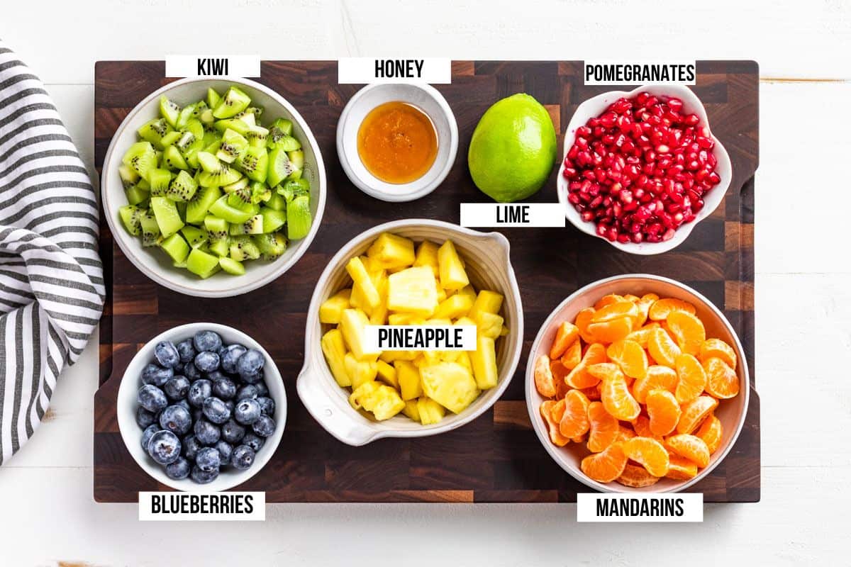 Bowls of cut up kiwi, pineapple, mandarin oranges, pomegranate arils, blueberries, honey and a lime on a wood board.