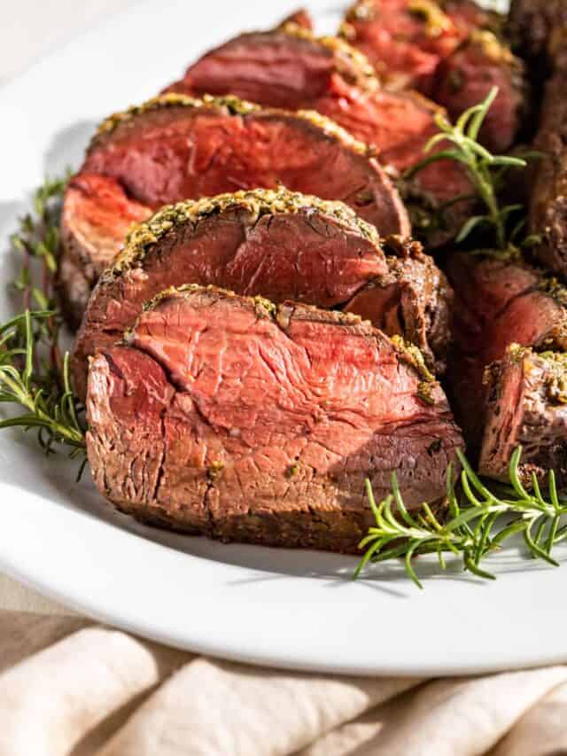 Sliced Roast Beef Tenderloin on a white plate surrounded by rosemary and thyme sprigs.