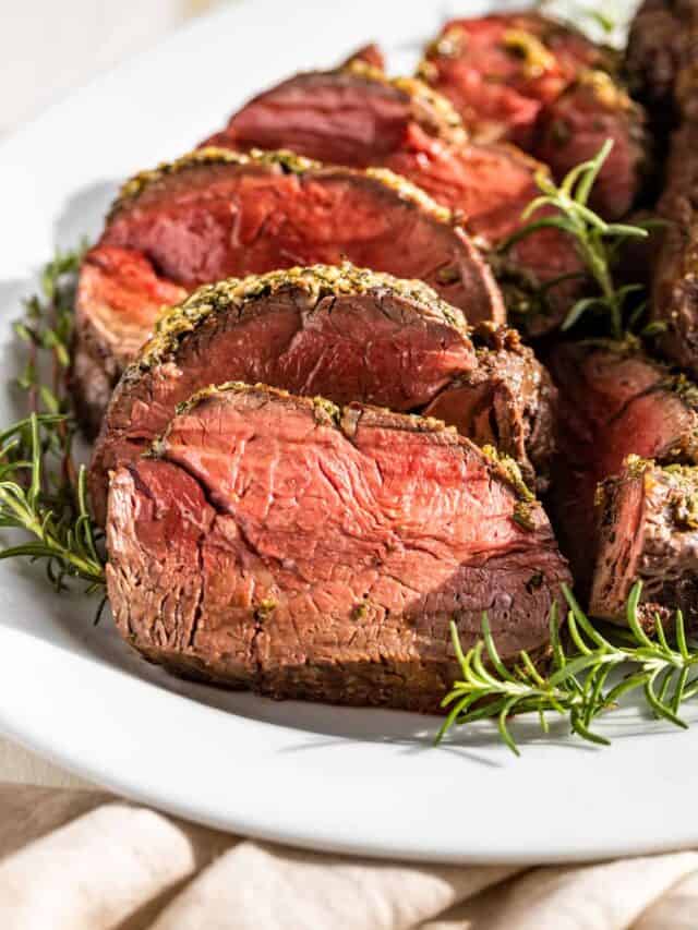 Sliced Roast Beef Tenderloin on a white plate surrounded by rosemary and thyme sprigs.