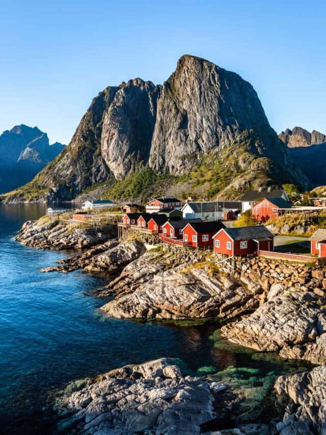 Red fishing houses on the edge of the water with mountains in the background.