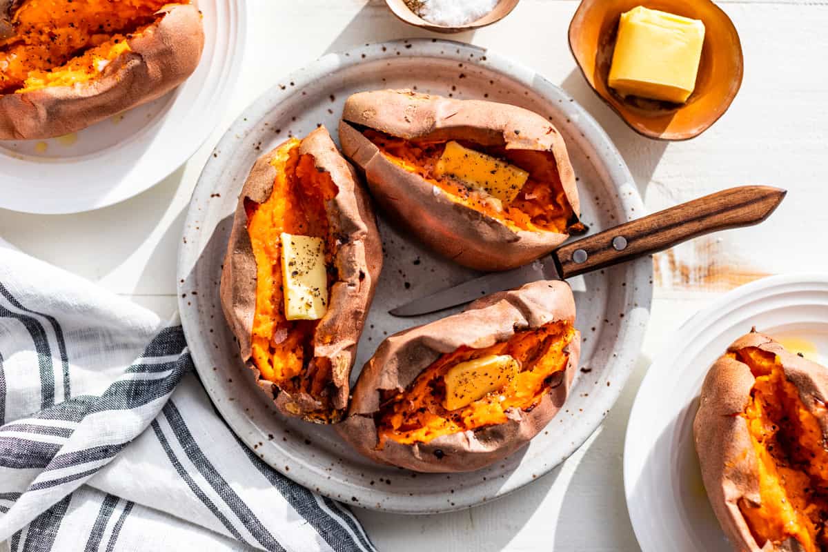 Baked sweet potatoes on a pottery plate split open with butter, sea salt, and pepper on the inside.