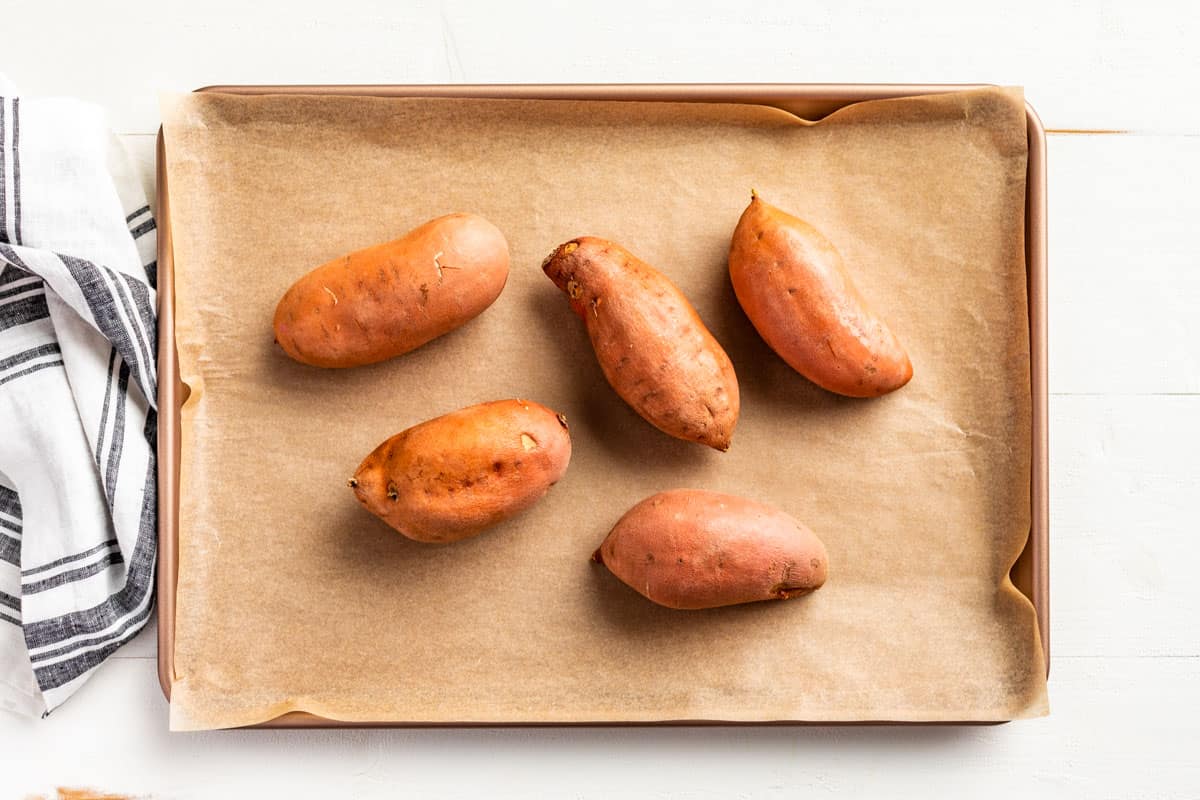 Washed sweet potatoes with slits made in them on a parchment lined baking sheet.