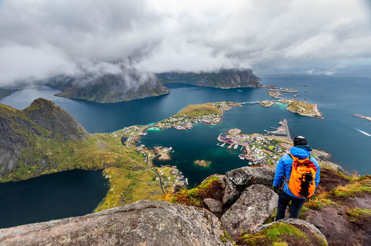 Man with an orange backpack standing on a mountain looking down on the city of Reine surrounded by ocean.