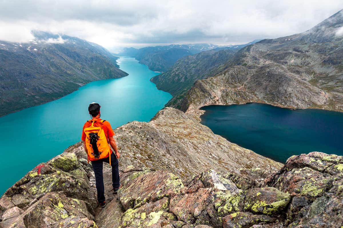 Man with an orange backpack standing on a rocky ridge between two glacially colored lakes.