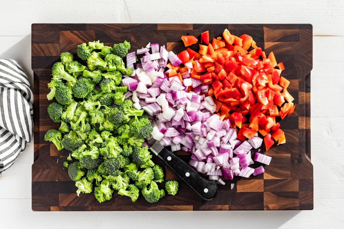 Chopped broccoli, diced red onion, and diced red bell pepper on a wood cutting board.