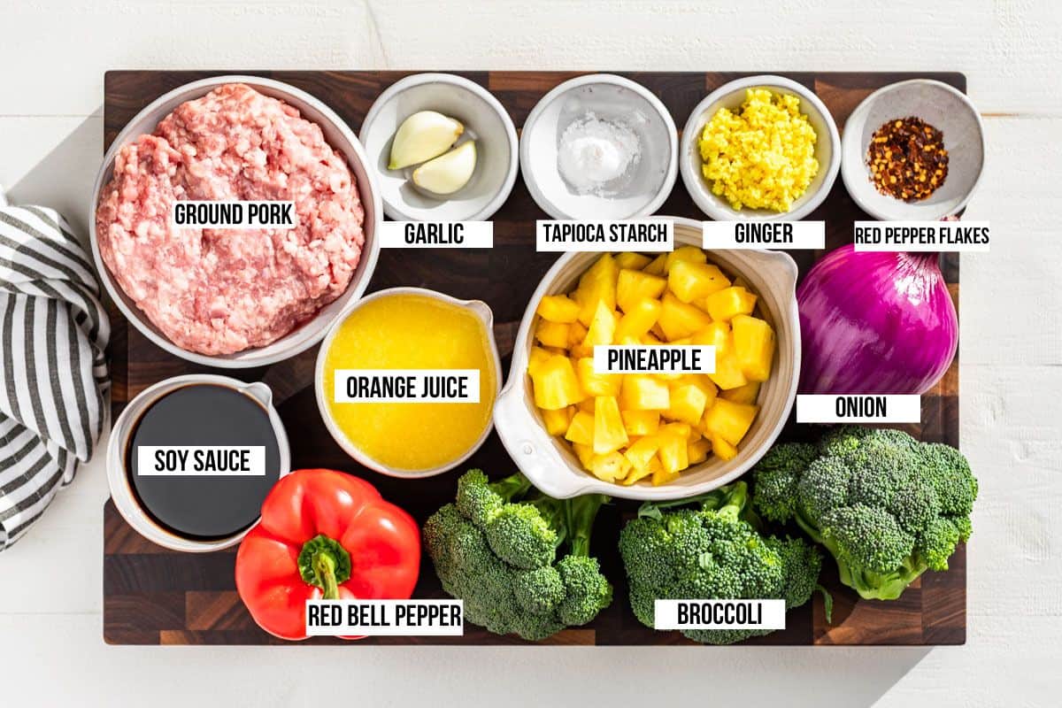 All the ingredients for Ground Pork Stir Fry measured out on a wood cutting board, ground pork, pineapple chunks, bell pepper, onion, broccoli, ginger, garlic, soy sauce, and orange juice.