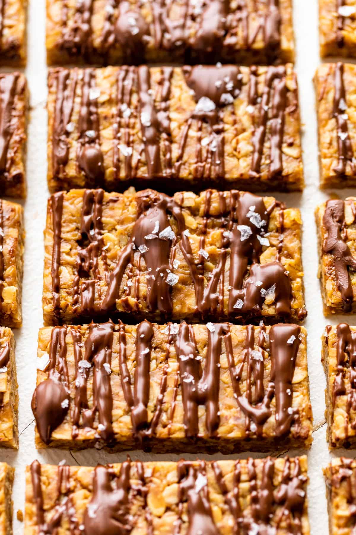 Side view of a row of Peanut Butter Granola Bars drizzled with melted chocolate and sprinkled with flaked sea salt.