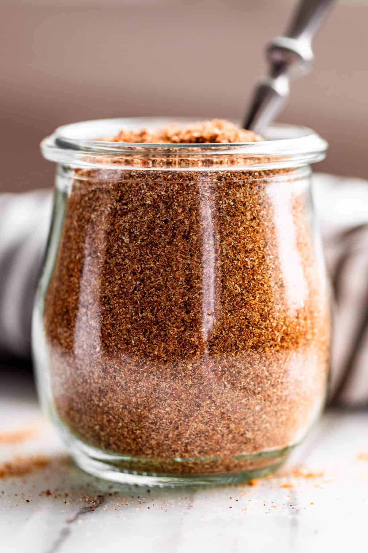 A clear glass jar of Taco Seasoning with a silver spoon in it and a light brown background.