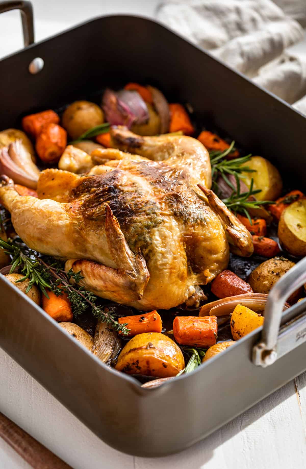 A whole roast chicken in a roaster surrounded with carrots, potatoes, and onions.