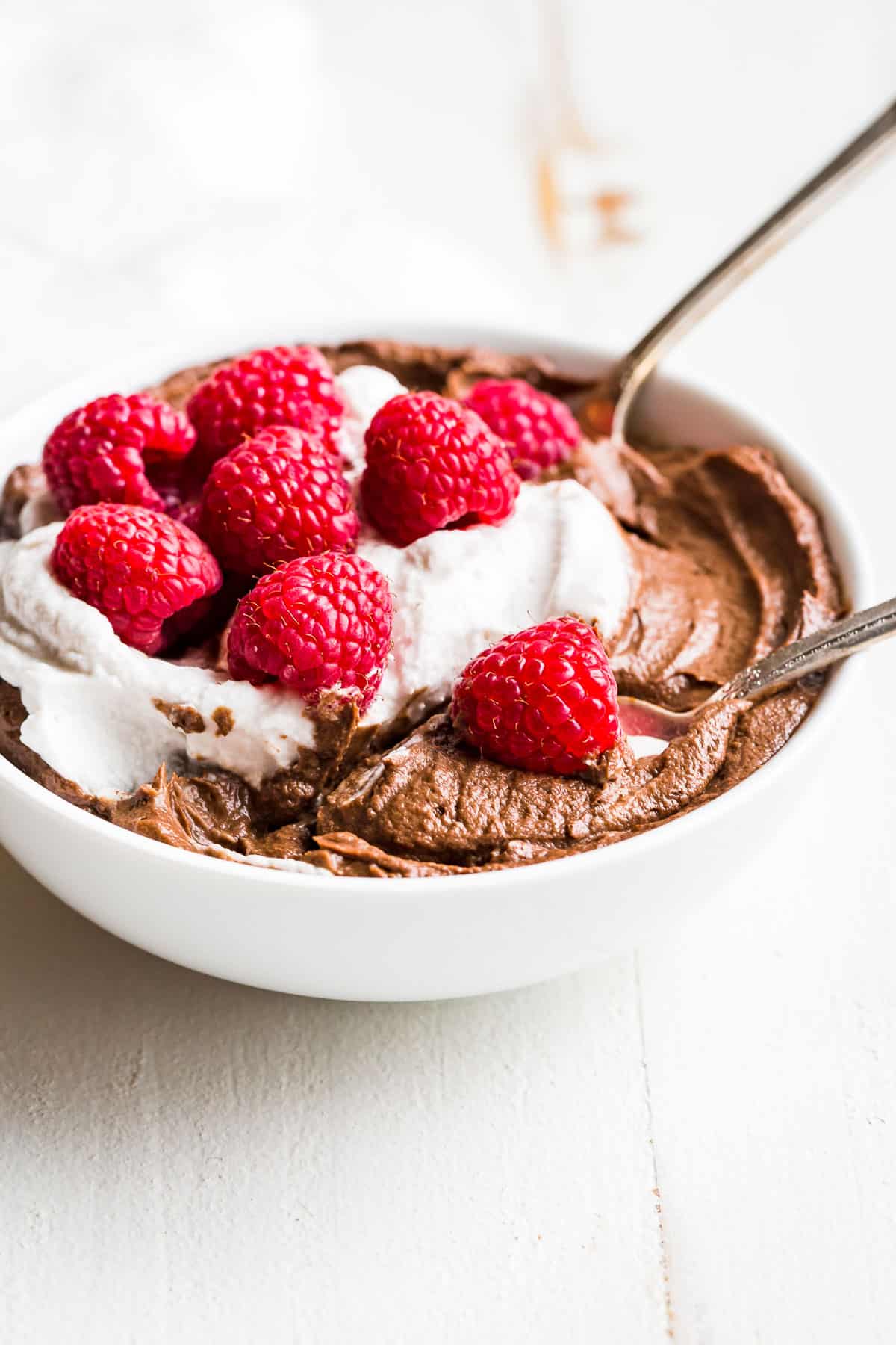 Side view of Avocado Chocolate Mousse in a white bowl topped with whipped cream and raspberries.