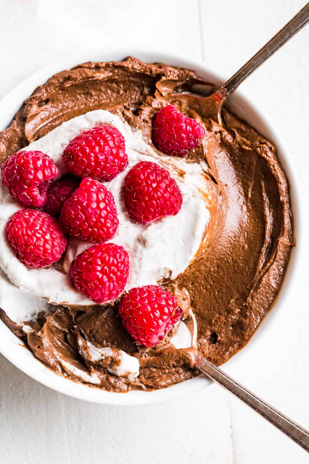 Straight down view of Avocado Chocolate Mousse in a white bowl topped with whipped cream and raspberries with 2 spoons in it.