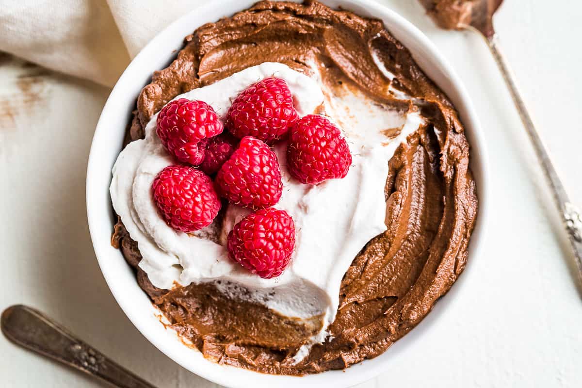 Straight down view of Avocado Chocolate Mousse topped with whipped cream and raspberries in a white bowl with 2 spoons on the side.