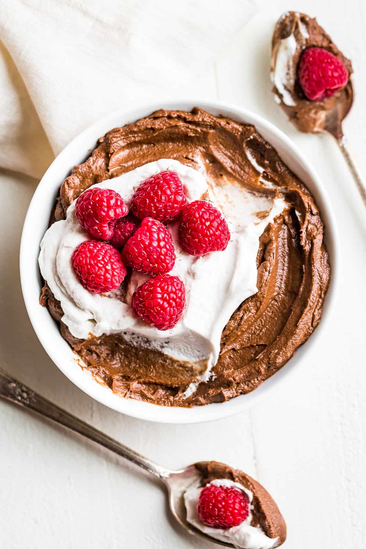 Straight down view of Avocado Chocolate Mousse in a white bowl topped with whipped cream and raspberries with 2 spoons scooping out 2 bites on the side of the bowl.